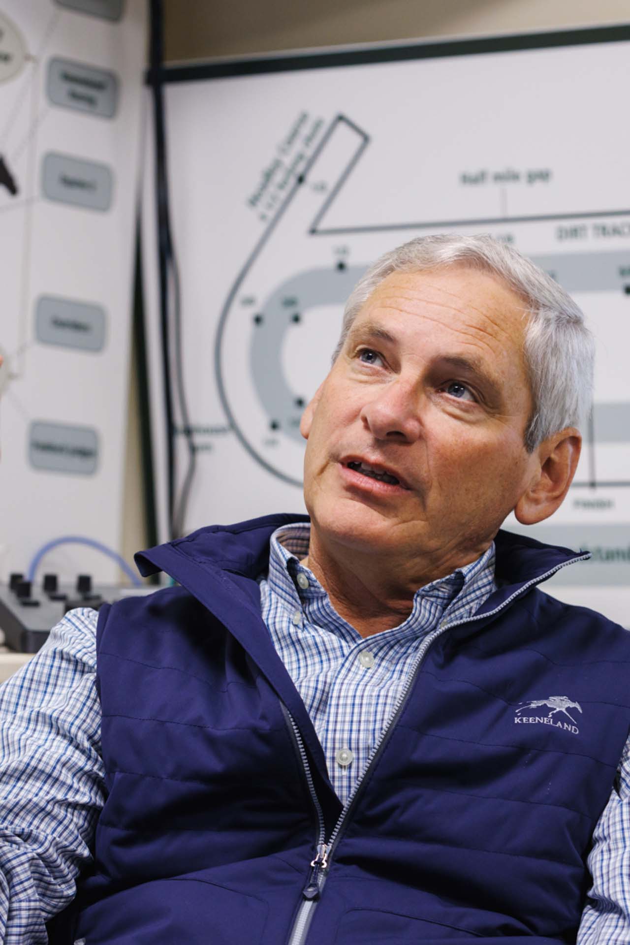 An up-close photo of Dr. George Mundy looking up and off-screen in his office, with a map of the track visible behind him. He is an older White man with short silver hair. He is wearing a navy Horsesrace-brand vest over a white and blue checkered shirt.